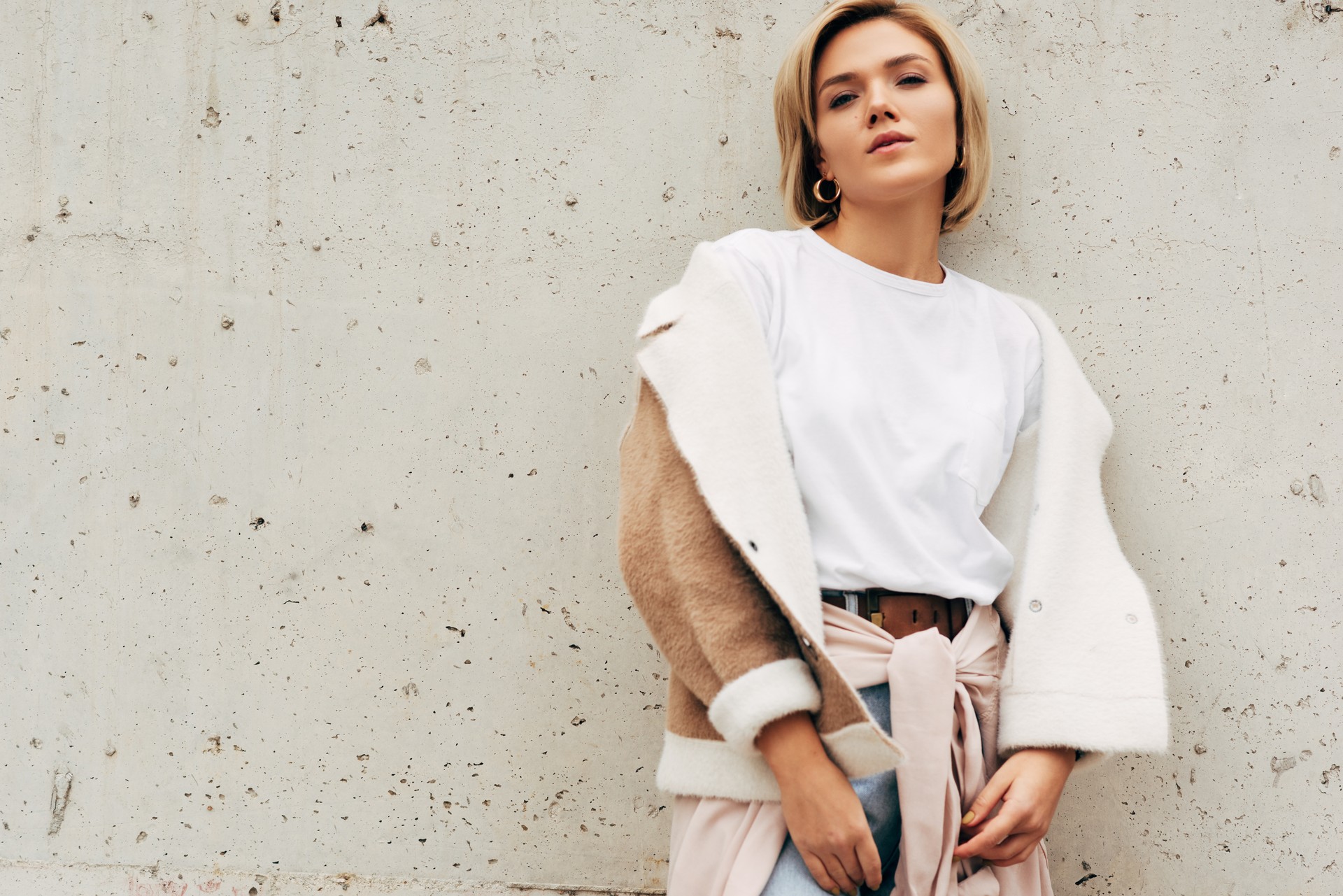 Image of stylish blonde young woman wearing white t-shirt and beige jacket, smiling and looking to the camera. Gorgeous female on concrete white wall in the city street, during waiting her boyfriend